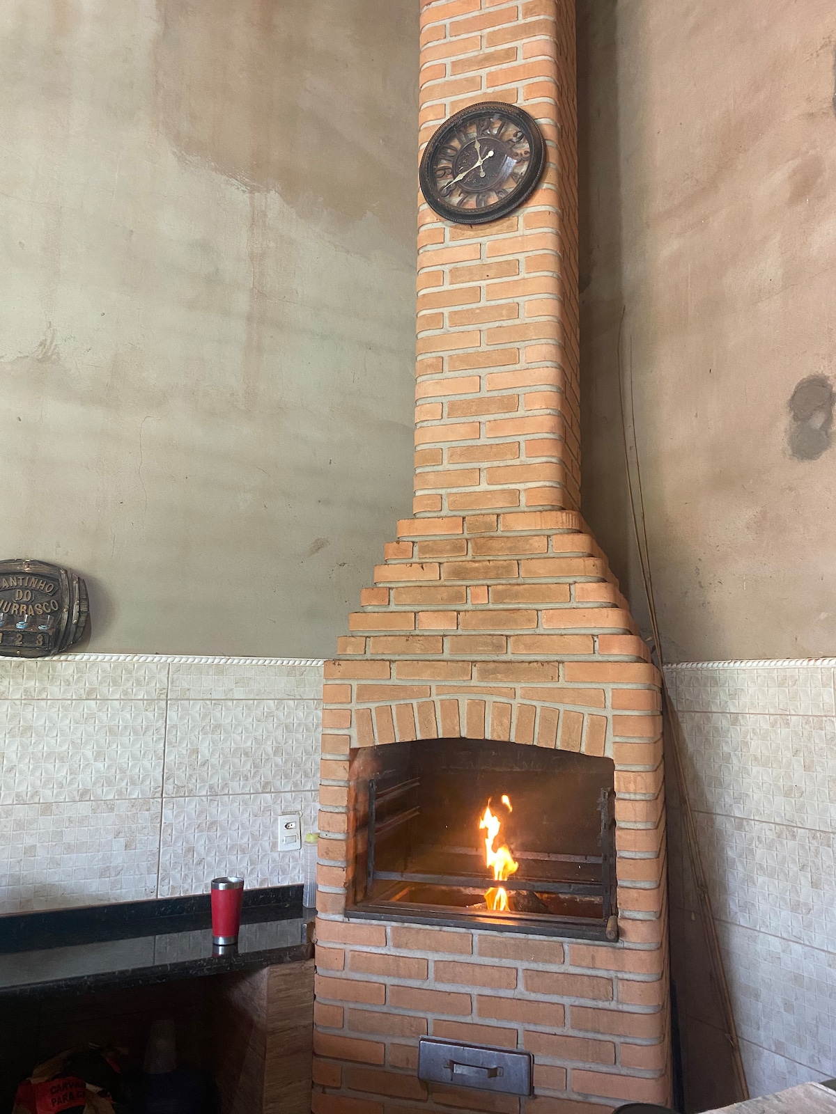 A tall brick fireplace is featured, with a clock mounted above its chimney. The flames from the fire are visible within the wood-burning area, providing warmth. The surrounding area includes a dark countertop, contributing to a homey cooking space.