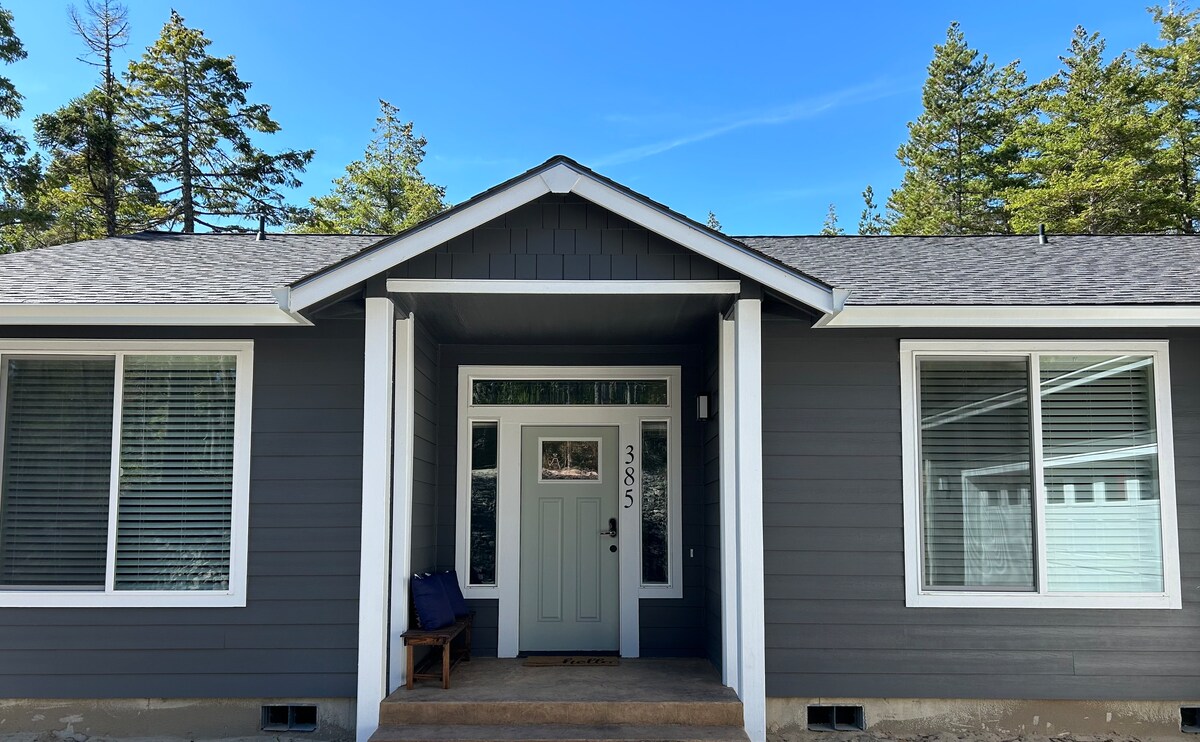 The entrance of the property features a welcoming front door framed by two large windows. A small wooden chair is placed to the left of the entryway, while tall trees are visible in the background under a clear blue sky.