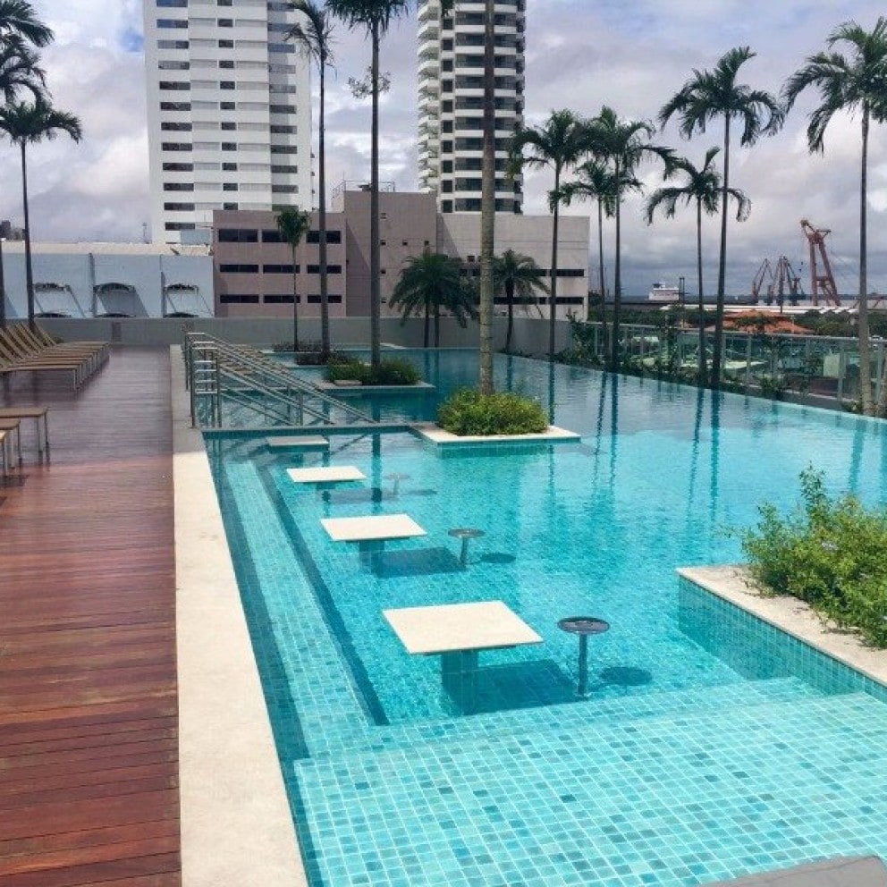 A modern pool area features clear blue water surrounded by palm trees and wooden lounge chairs. Stone platforms are positioned within the pool, creating a unique visual effect. The backdrop includes tall buildings and a hint of greenery, contributing to a relaxing outdoor environment.
