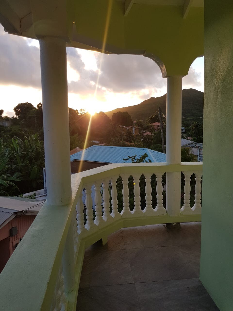 A balcony with ornate white railings overlooks a landscape at sunset. The warm hues of the horizon contrast with the lush green hills and rooftops below, creating a serene backdrop for relaxation. Soft light filters through the scene, enhancing the peaceful atmosphere.