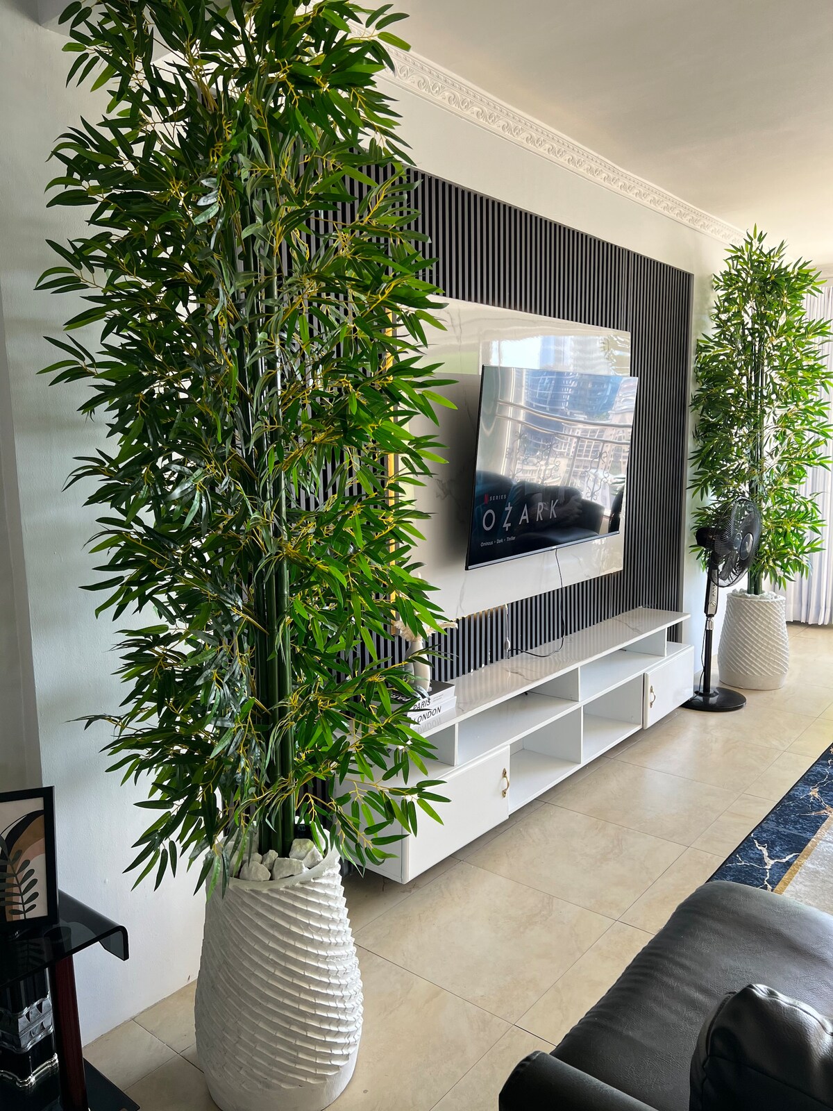 A modern living area features a wall-mounted television above a sleek entertainment unit. Two tall, decorative plants in white planters provide greenery on either side. A fan stands nearby, and natural light filters through a window, illuminating the beige tiled flooring.