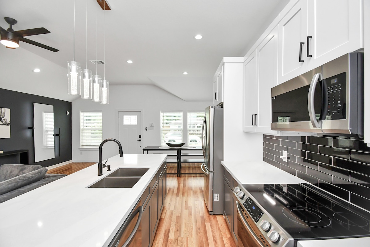 The kitchen features modern stainless-steel appliances, including a microwave and oven, set against sleek black tile. A large island with a dual sink is complemented by overhead pendant lighting. Natural light fills the space through multiple windows, contributing to the open layout.