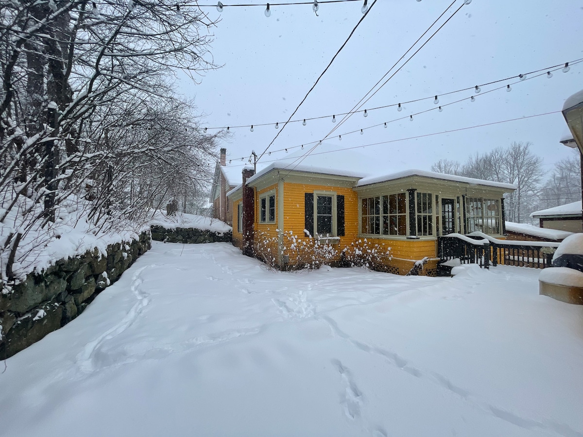 A quaint yellow cottage is enveloped in fresh snow, with pathways leading to the entrance. The enclosed porch, adorned with large windows, offers a view of the winter landscape. String lights hang overhead, adding subtle charm to the serene outdoor setting.