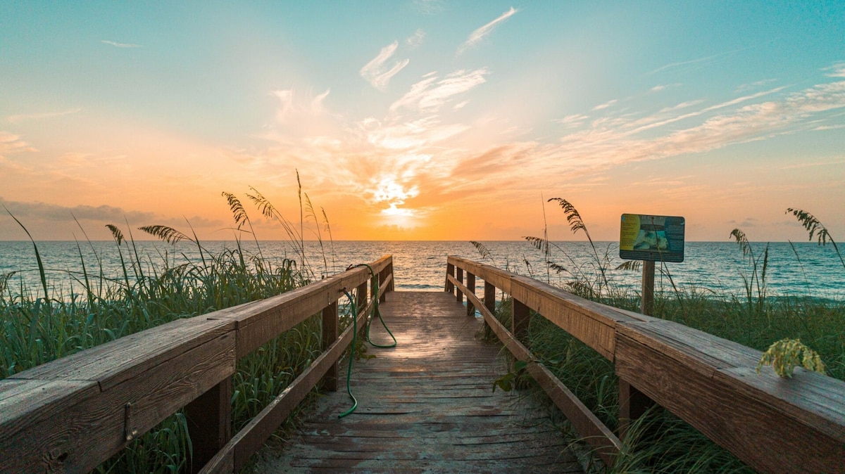 A wooden boardwalk extends towards the horizon, leading through sea oats to a sunset over the ocean. Soft colors of orange and blue blend in the sky, while gentle waves meet the sandy shore in the distance.