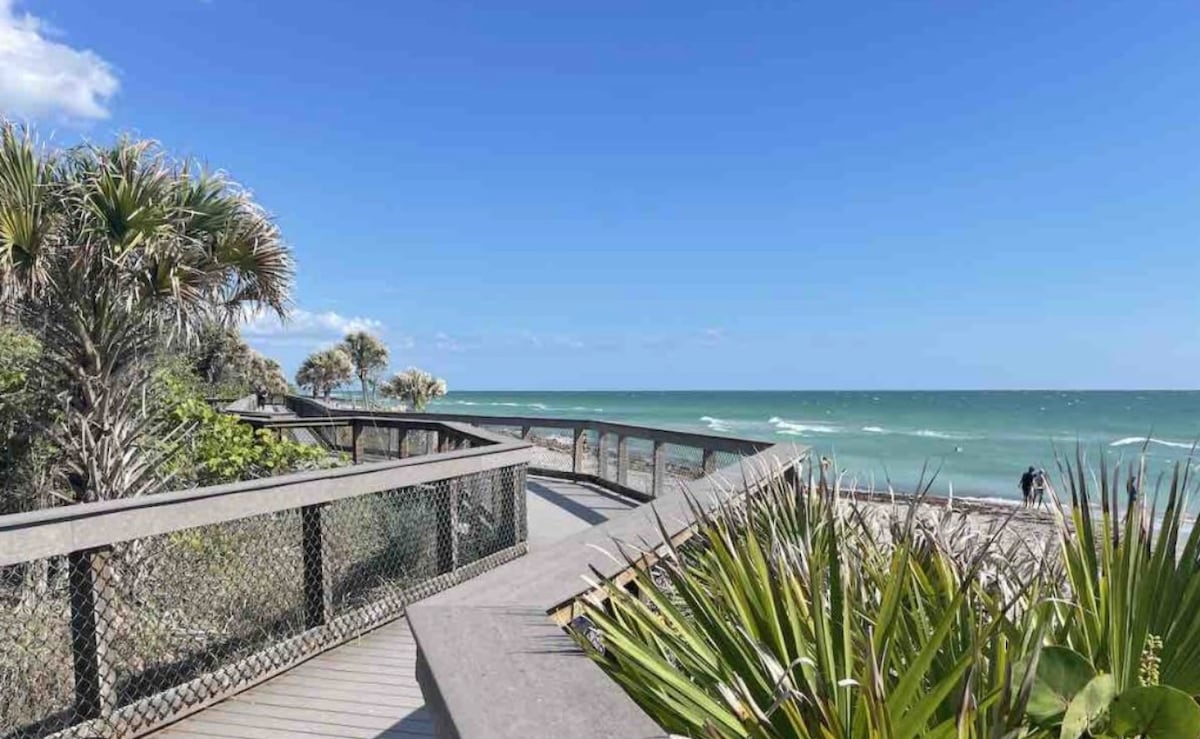 A wooden boardwalk leads toward a sandy beach, framed by palm trees and coastal vegetation. The ocean stretches out in the distance, with gentle waves rolling in under a clear blue sky. Sunlight brightens the scene, inviting relaxation and enjoyment of the natural surroundings.