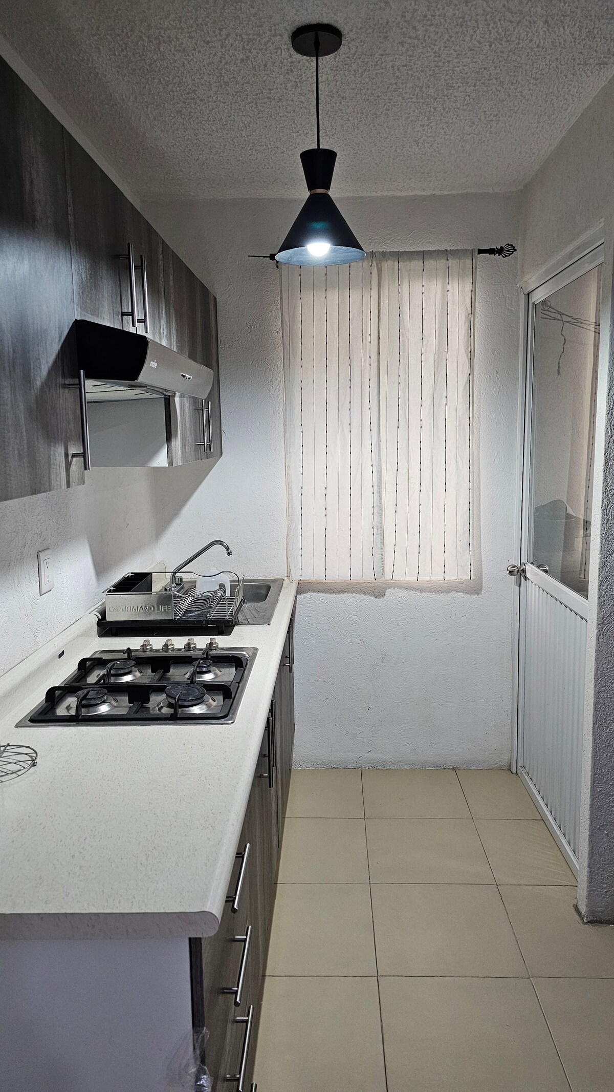 A modern kitchen is equipped with a gas stove, sleek countertops, and ample cabinetry. Natural light filters through a sheer curtain covering a window, and a door leads to an external area. A pendant light hangs above the workspace, illuminating the cooking area.