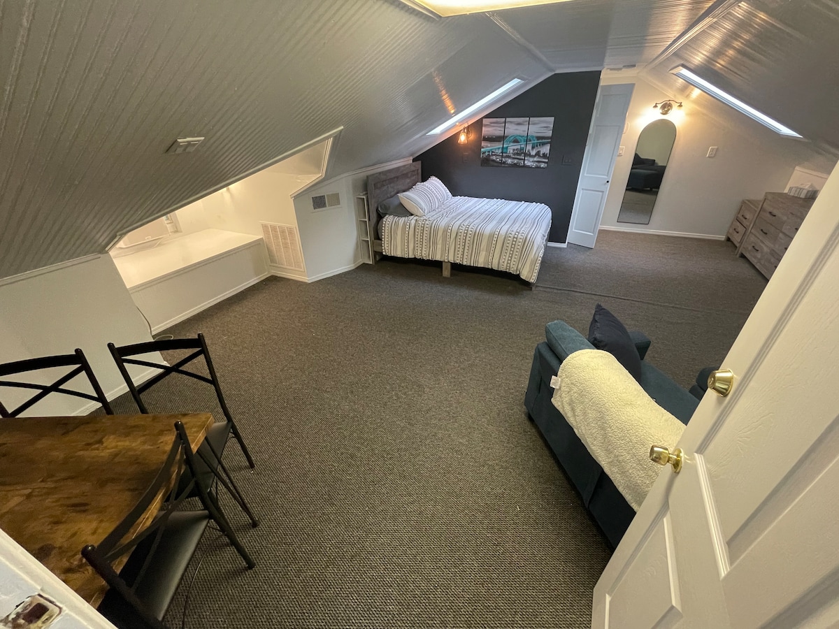 The spacious room features a light-colored queen bed with striped bedding, positioned under the slanted ceiling. A cozy couch is visible, alongside a wooden dining table and chairs, creating an inviting space. A door leads to an adjoining area, enhancing accessibility.