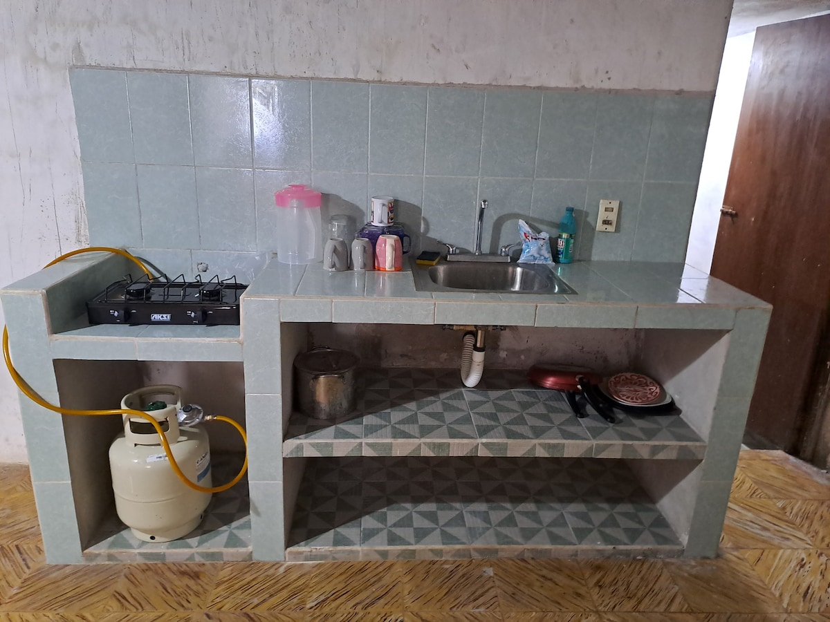 A practical kitchen area features tiled surfaces in soft blue tones. A sink, a gas stove, and various cooking utensils are visible. Underneath, open shelving provides space for pots and dishes, while a gas cylinder sits beside the counter.
