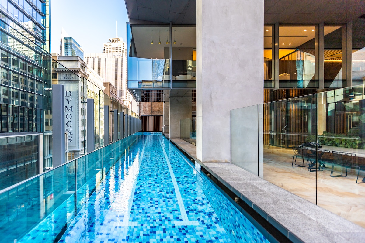 A heated horizontal pool is seen extending along the building's edge, featuring a mosaic tile design in varying shades of blue. Floor-to-ceiling glass panels offer views of the surrounding cityscape, including modern skyscrapers and outdoor seating areas in proximity.