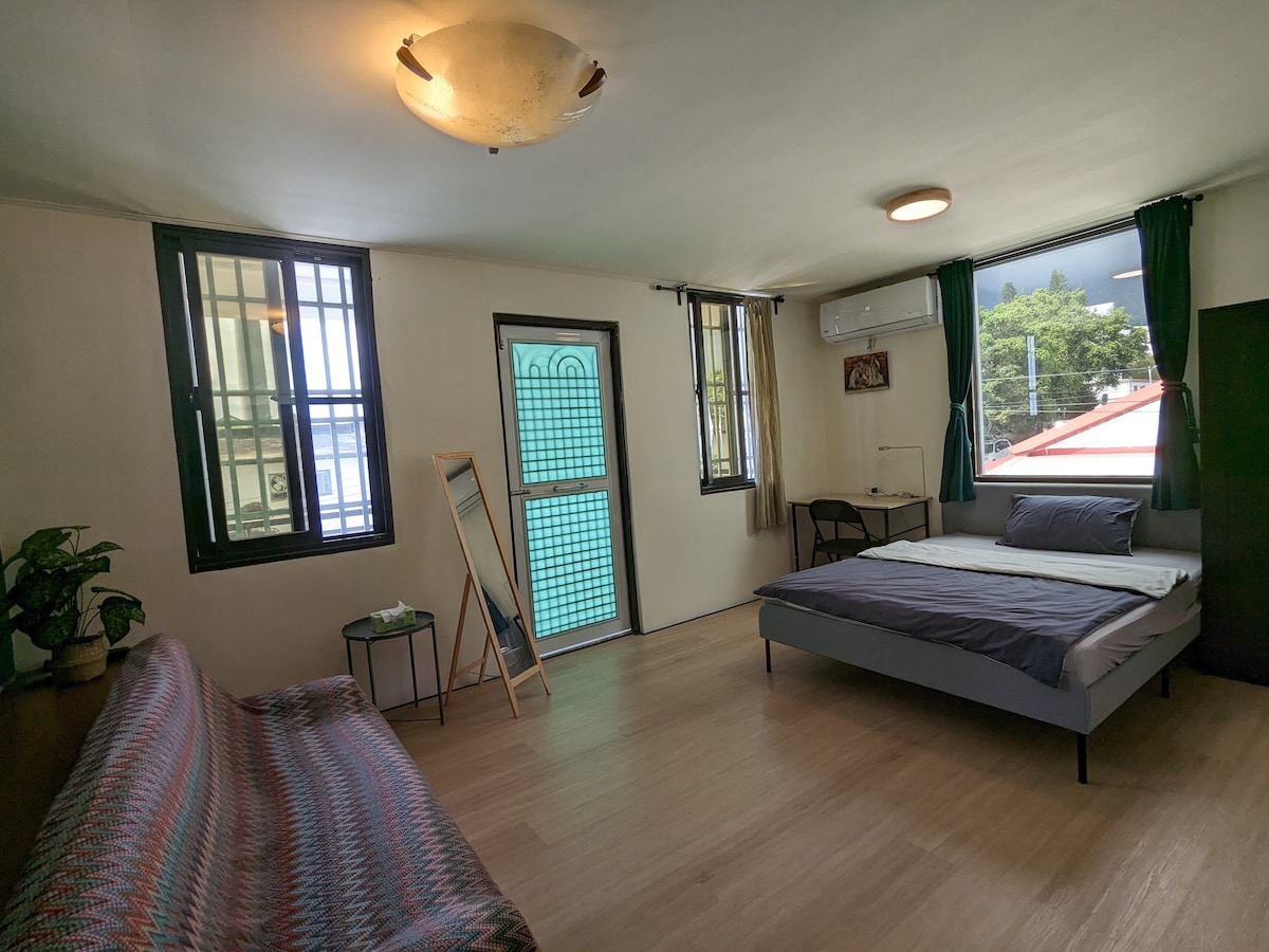 A spacious room features a double bed with clean linens and a sofa bed, complemented by a desk. Two windows provide natural light, while a door leads to a balcony. A plant adds a touch of greenery, and a full-length mirror is positioned against the wall.