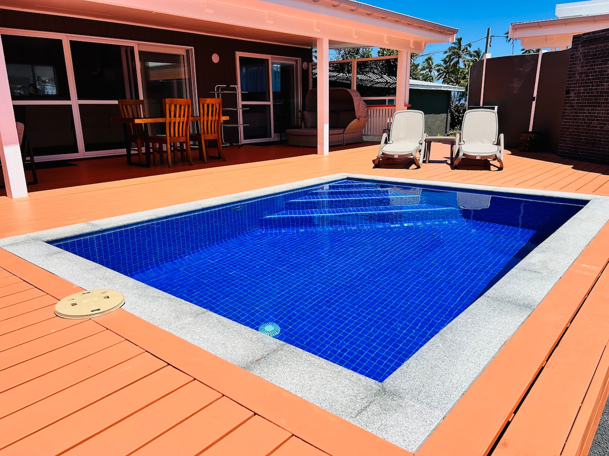 A private pool is surrounded by a spacious deck, featuring padded lounge chairs for relaxation. The bright blue water of the pool contrasts with the warm orange tones of the decking, inviting moments of leisure outdoors in a sunlit area.