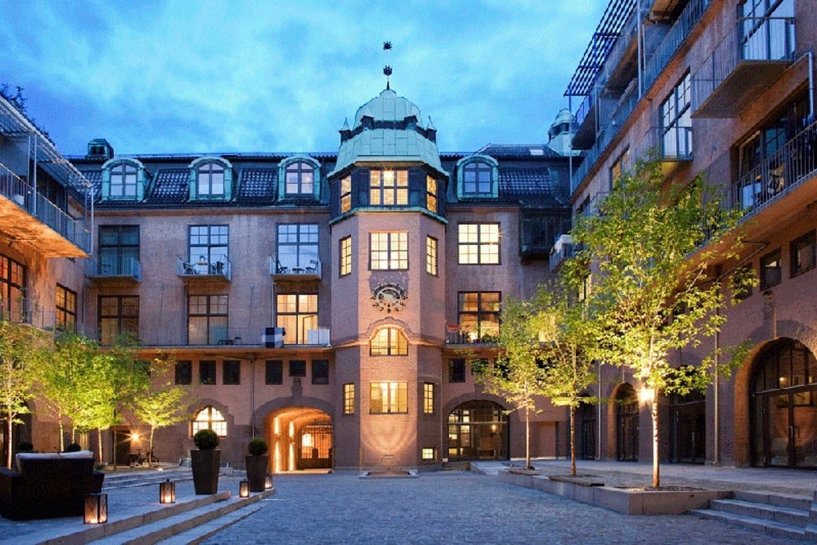 An inviting courtyard is showcased, featuring a historic building with a distinctive turret and large windows. Soft lighting enhances the surroundings, with neatly trimmed trees lining the stone pathway. Balconies adorned with plants overlook the area, creating a serene and welcoming atmosphere.