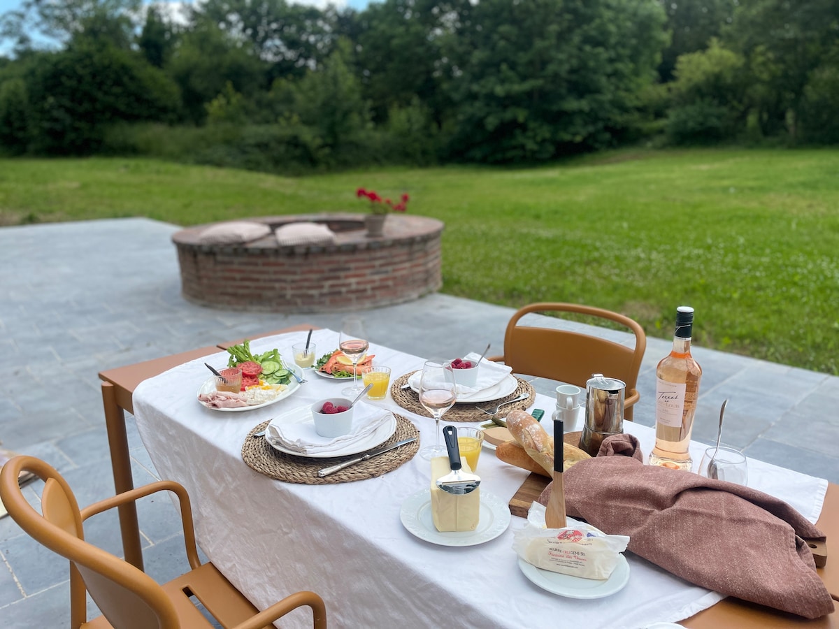 A dining setup is displayed on a spacious terrace, featuring a white tablecloth and neatly arranged dishes. Plates of fresh salad, bread, and desserts are complemented by wine glasses. In the background, a green expanse is visible, accented by trees and a brick fire pit.