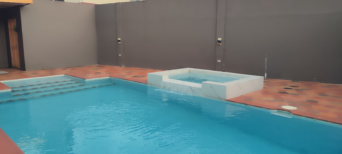 A tranquil pool area is featured, showcasing a spacious swimming pool complemented by a separate hot tub. Surrounding the pool is a tiled deck, with a neutral-colored wall in the background offering a serene ambiance.