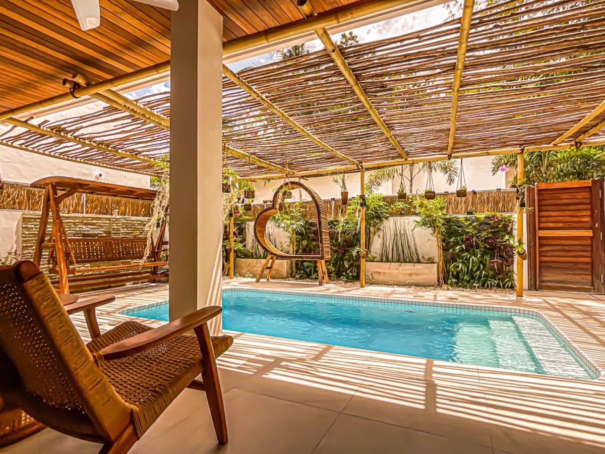 A private pool area is presented with a spacious swimming pool surrounded by greenery. A covered patio offers shaded seating, featuring a swing chair and a woven lounge chair. Natural light filters through bamboo overhead, creating a serene environment for relaxation.