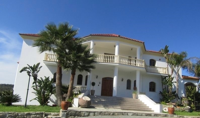 The villa's elegant facade features a symmetrical design complemented by a terracotta roof. Expansive stairs lead to the main entrance, framed by decorative pots and lush green plants. Large balcony railings overlook the surroundings, highlighting the property's inviting outdoor appeal.