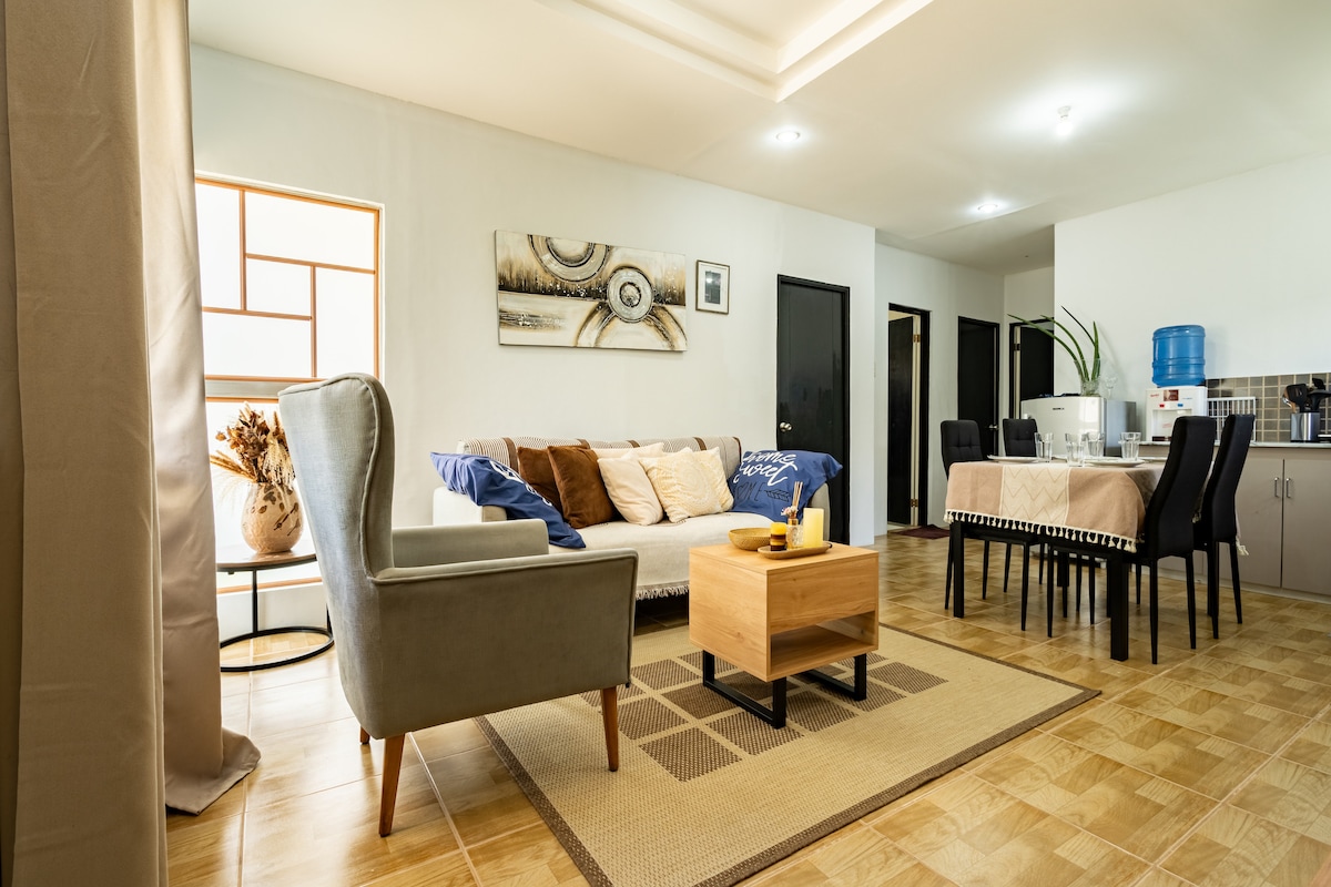A light-filled living area is furnished with a comfortable sofa adorned with decorative cushions, accompanied by a stylish armchair. A wooden coffee table sits on a textured rug, and a dining table with chairs is visible in the background. Minimalist wall art adds a subtle touch.