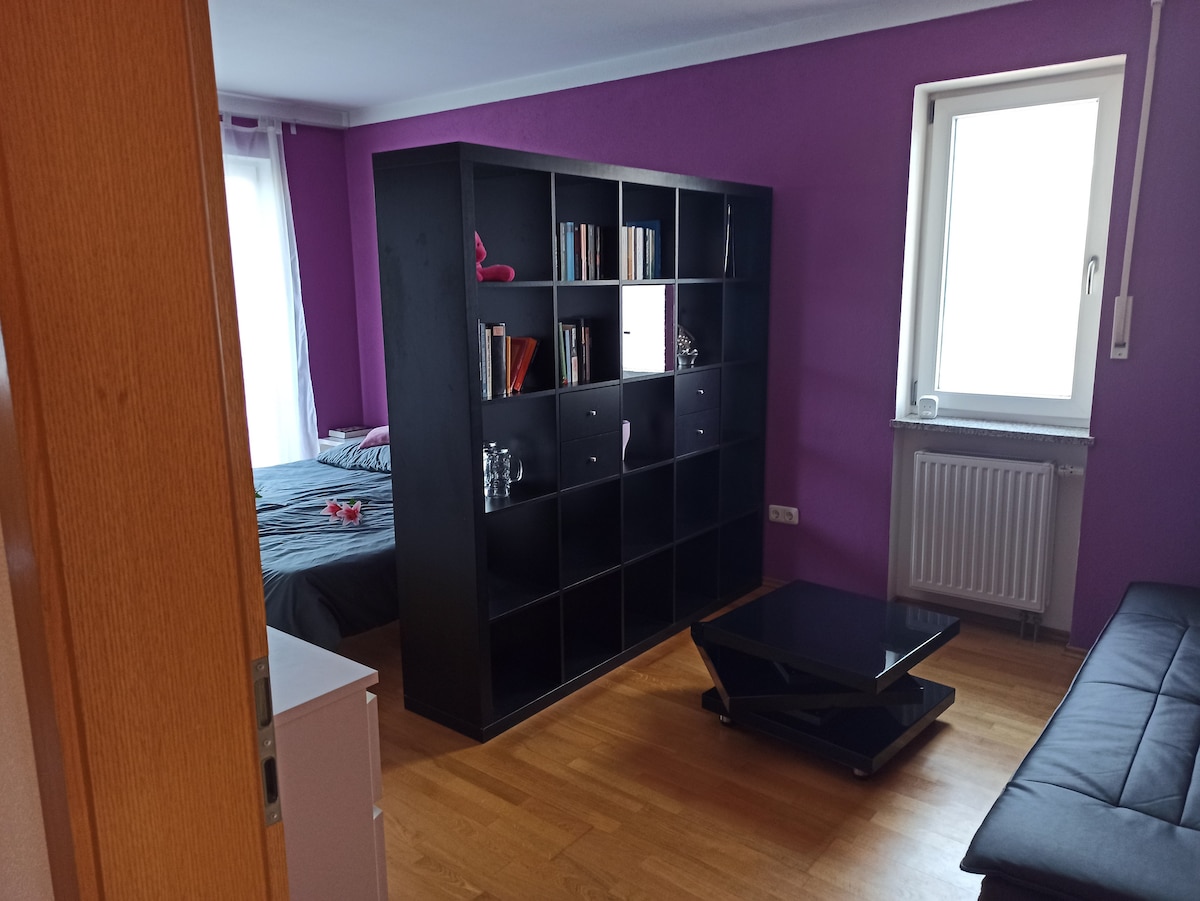 A bright room features a purple accent wall with a spacious bed visible opposite a black bookshelf filled with books and decorative items. A sleek black coffee table is positioned in front of a foldable couch. Natural light enters through a window, highlighting the hardwood flooring.