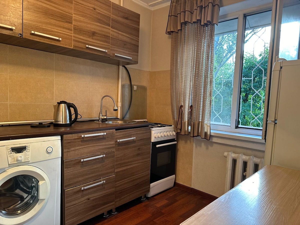 A functional kitchen area features wooden cabinetry and modern appliances. A kettle rests on the countertop beside a washing machine. An oven and stovetop are positioned next to a window that provides natural light, framed with patterned curtains.