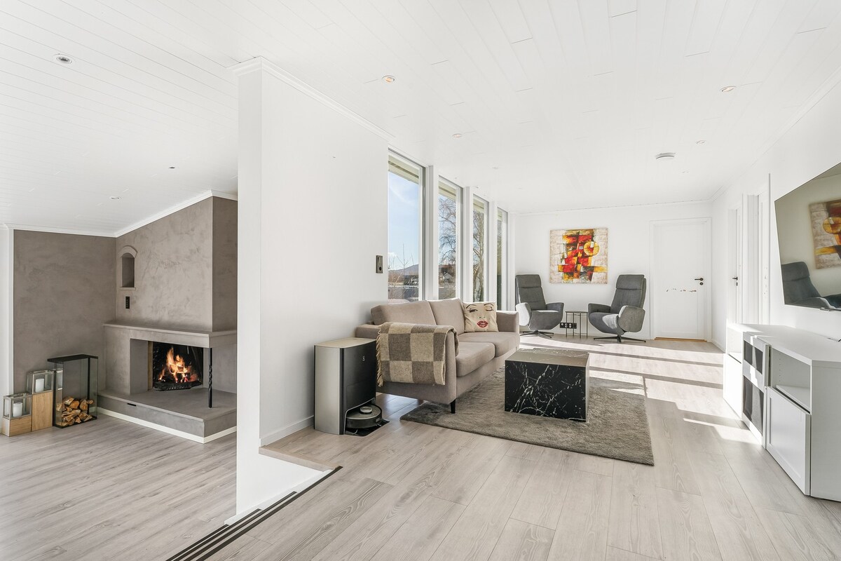 The living area showcases a neutral color palette with wooden flooring and ample natural light. A minimalist fireplace is positioned against a feature wall. Comfortable seating arrangements are evident, complemented by contemporary artwork and a stylish coffee table.