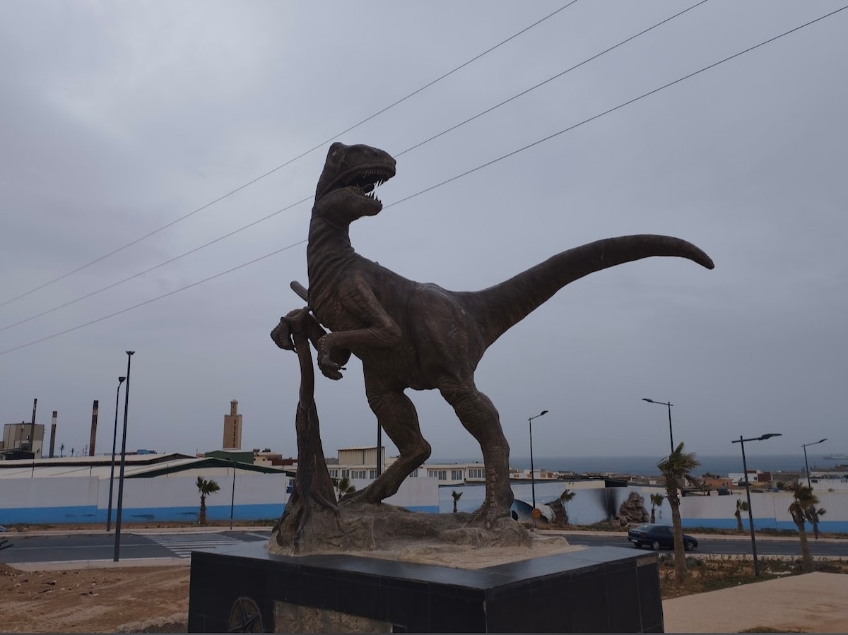 A detailed bronze statue of a dinosaur is positioned on a pedestal, showcasing the creature in an aggressive stance while clutching a smaller figure. The background features a cloudy sky and distant buildings, with streetlights lining the area.