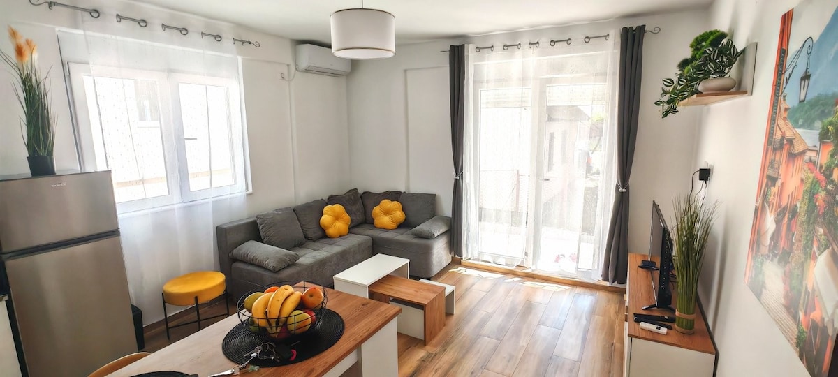 A living area features a comfortable gray seating arrangement with decorative yellow cushions. Sunlight filters through sheer curtains, illuminating a light wood floor. A small dining table with a fruit centerpiece and a television is visible, creating a cozy atmosphere for relaxation.