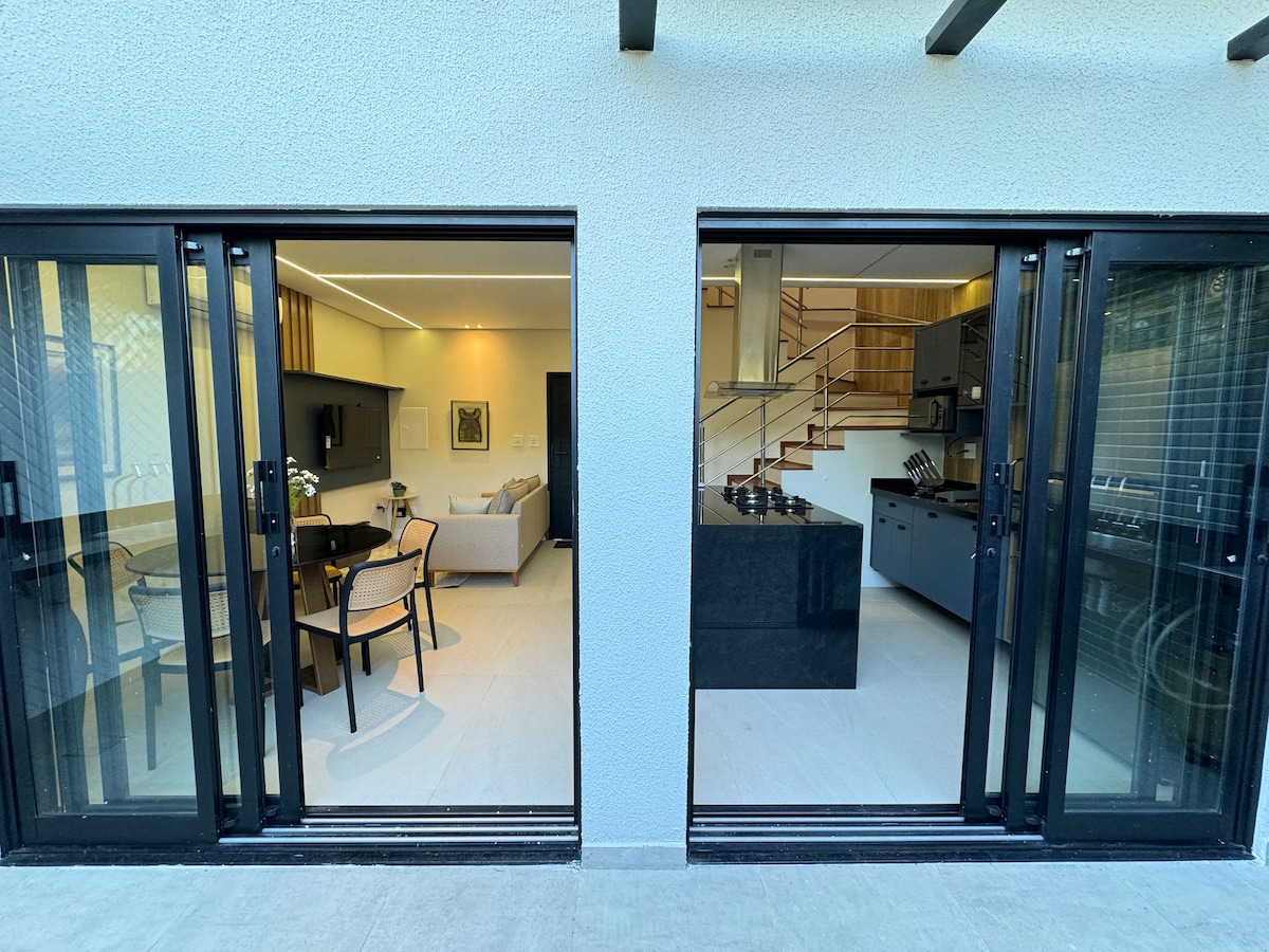 The image showcases an open space divided by large sliding glass doors. On one side, a dining area with a table and chairs is visible, while the other features a modern kitchen with a sleek black countertop and appliances. Natural light fills the area.
