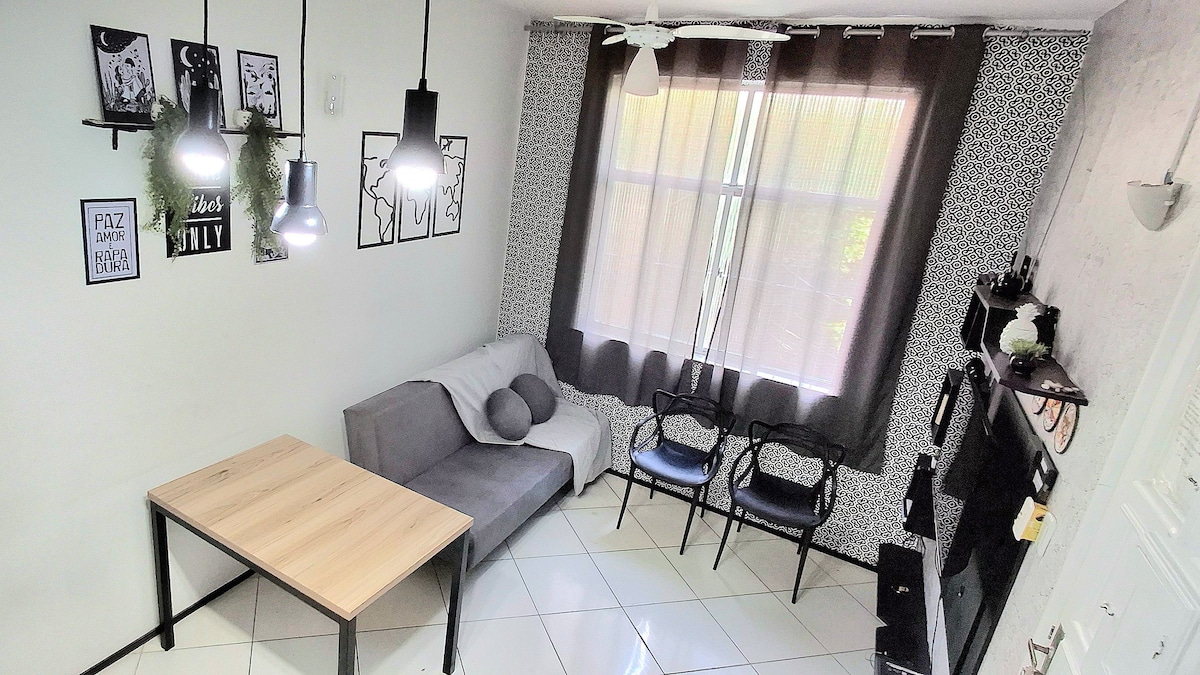 A light-filled living area features a gray sofa with cushions and a wooden table. Black chairs provide additional seating. Artwork is displayed on the wall, while large windows are dressed with dark curtains, allowing soft light to illuminate the space.