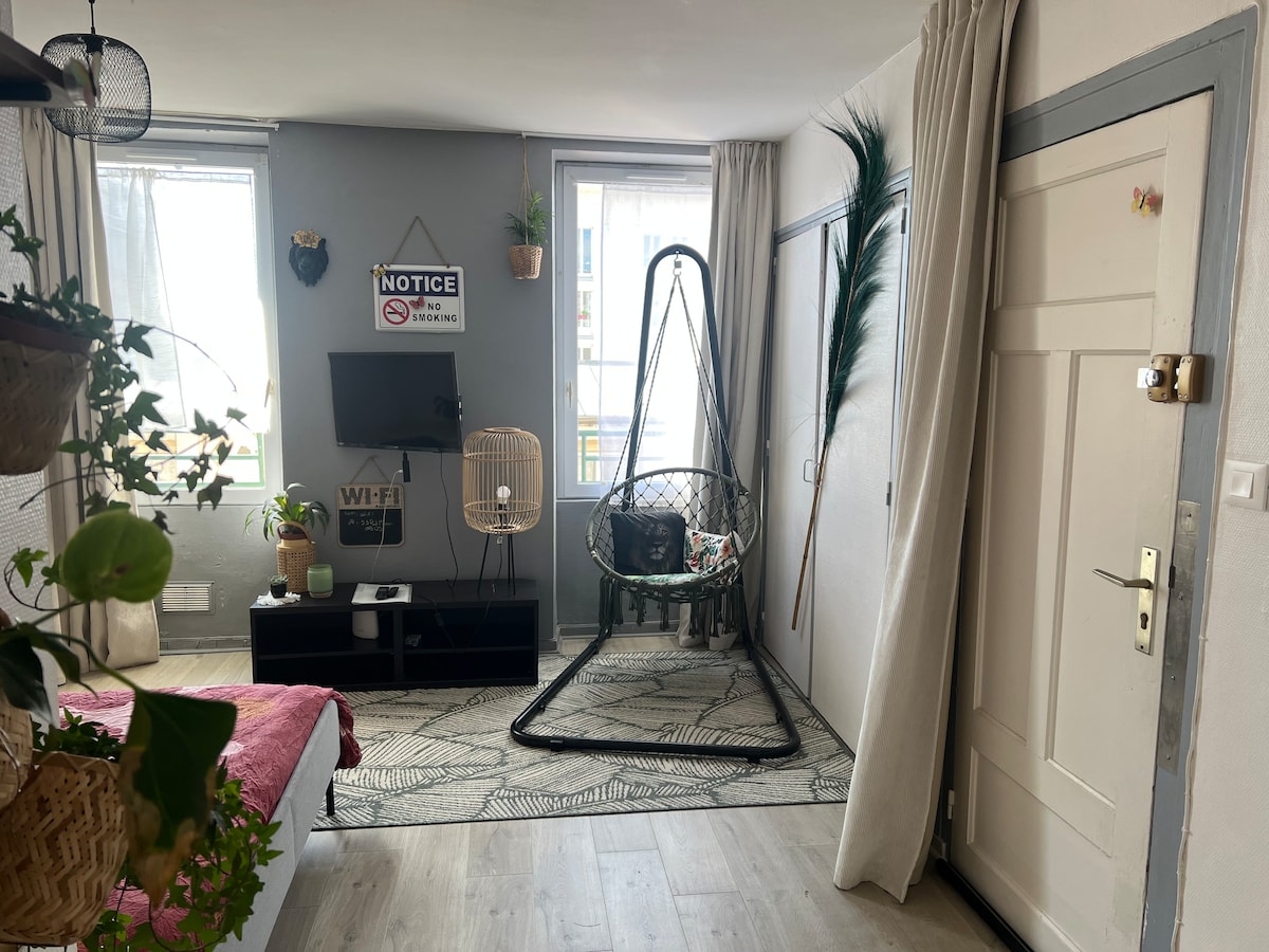 A cozy living space features a hanging chair in the corner, accompanied by a wall-mounted television. Natural light enters through two windows, brightening the room. A decorative rug covers the floor, while plants add a touch of greenery to the inviting environment.