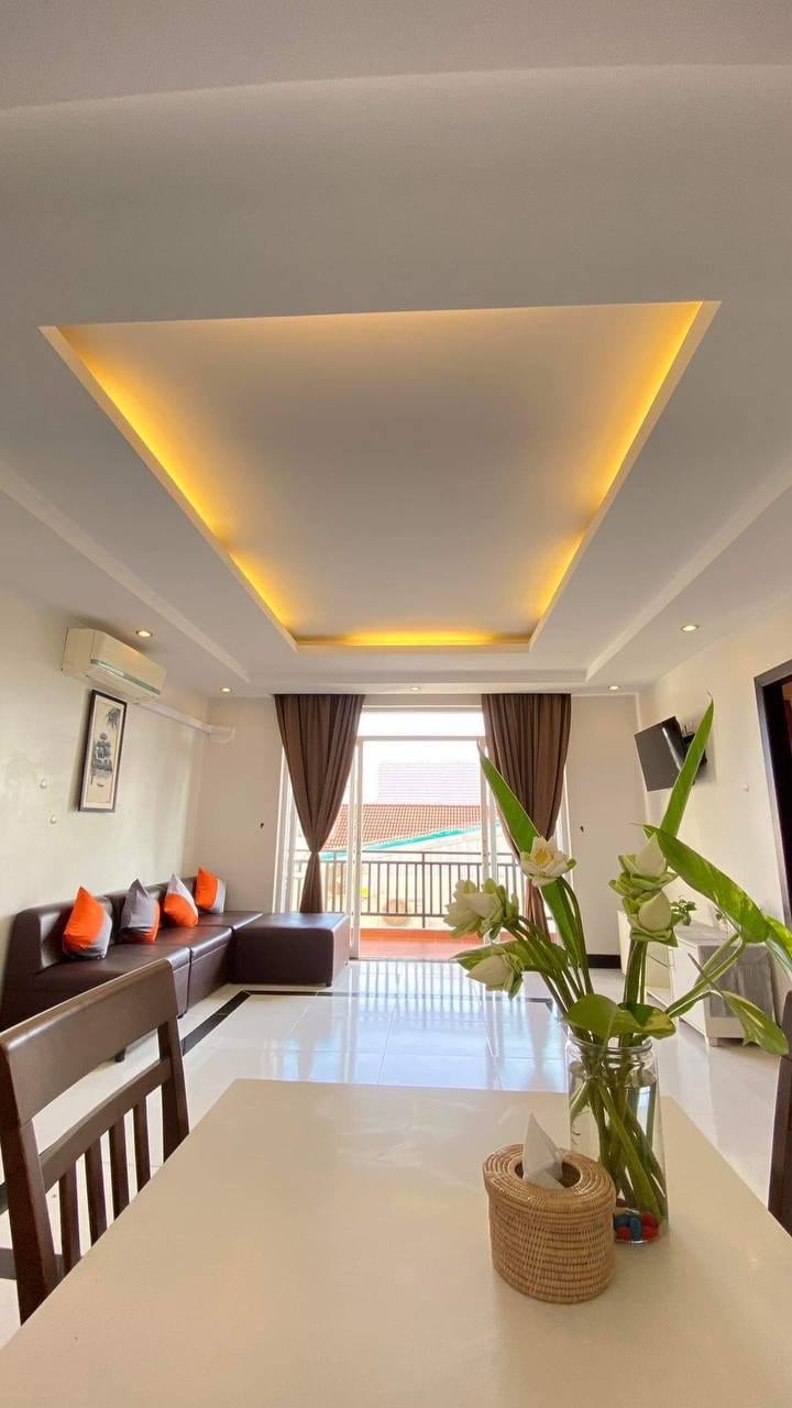 A modern living space features a sleek ceiling with integrated lighting above a cozy seating area, with a brown leather couch paired with brightly colored cushions. A dining table is positioned near a window, allowing natural light to illuminate the room.