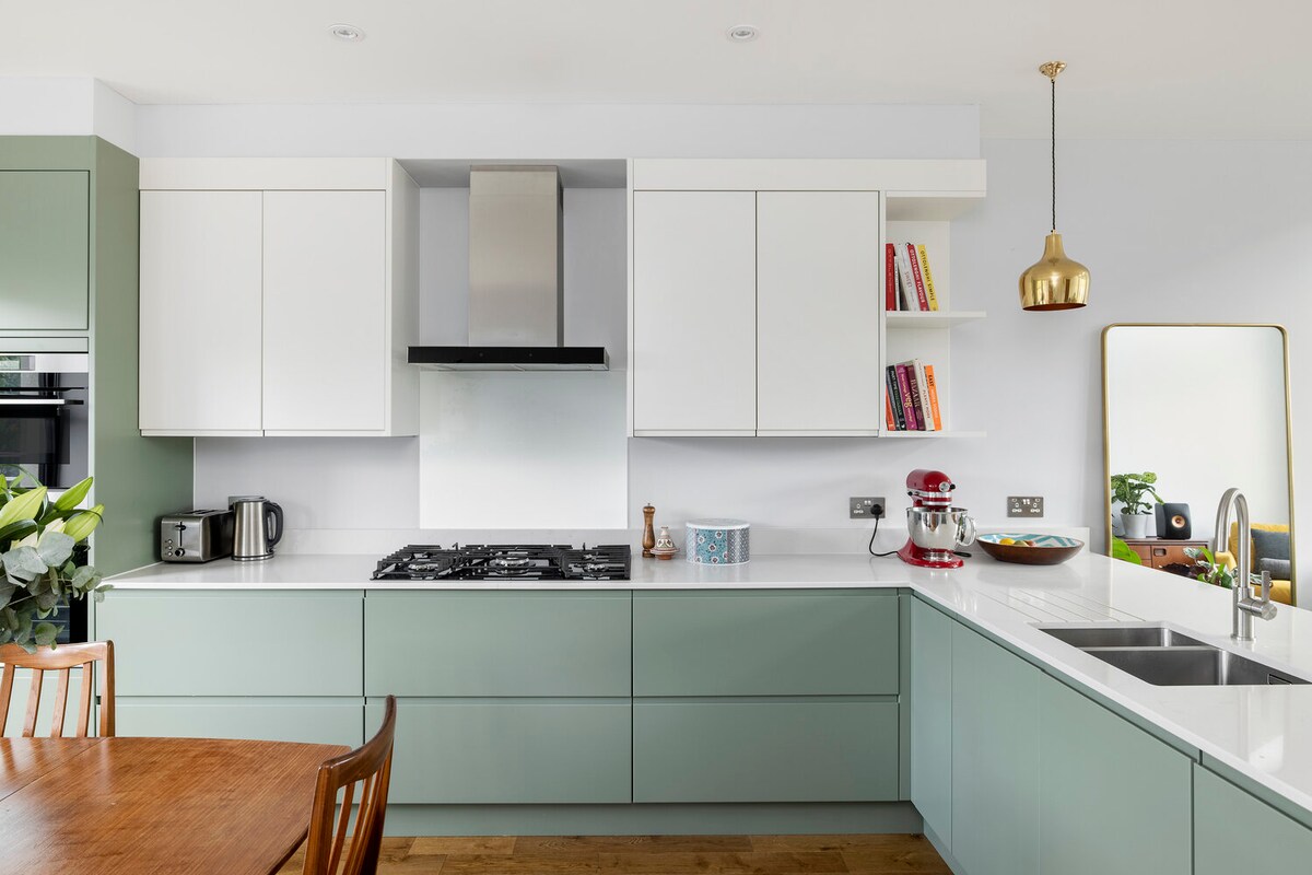 A modern kitchen features light green cabinetry paired with white upper cabinets, an integrated oven and gas hob. A breakfast bar is complemented by wooden dining chairs. A stylish mirror and potted plants add natural elements to the space, enhancing its inviting feel.