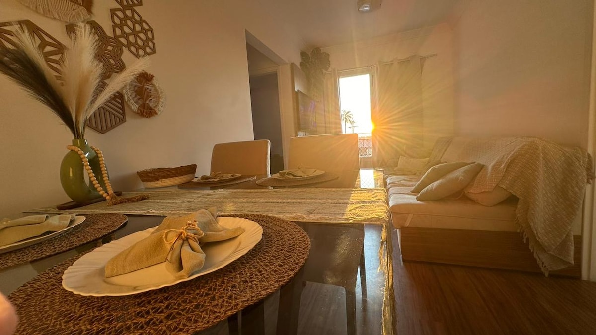 A dining area is presented with a woven table runner and plates elegantly arranged. Sunlight filters through the window, creating a warm ambiance. A comfortable sofa is visible in the background, complemented by decorative elements on the walls.