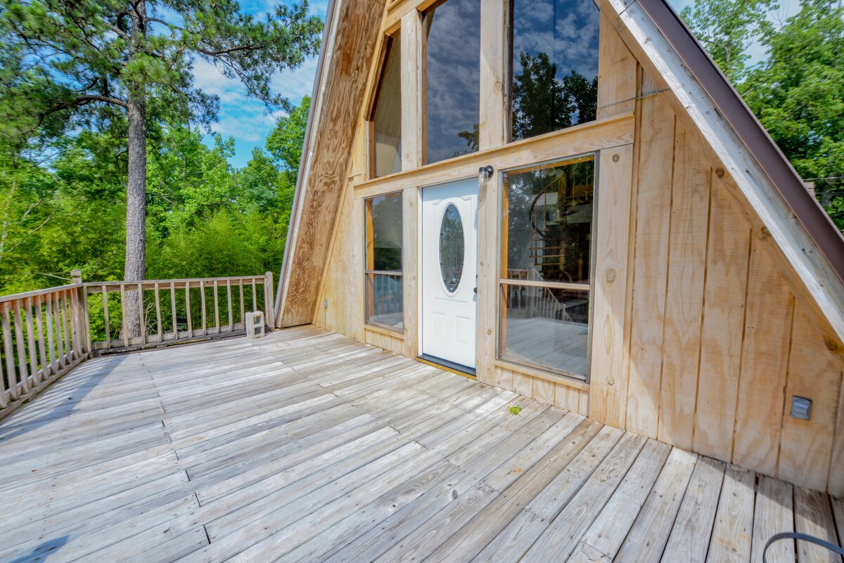 A spacious wooden deck extends from the entrance of the A-frame house, featuring large glass windows that provide plentiful natural light. The wood siding of the structure complements the surrounding greenery, creating a serene outdoor space.