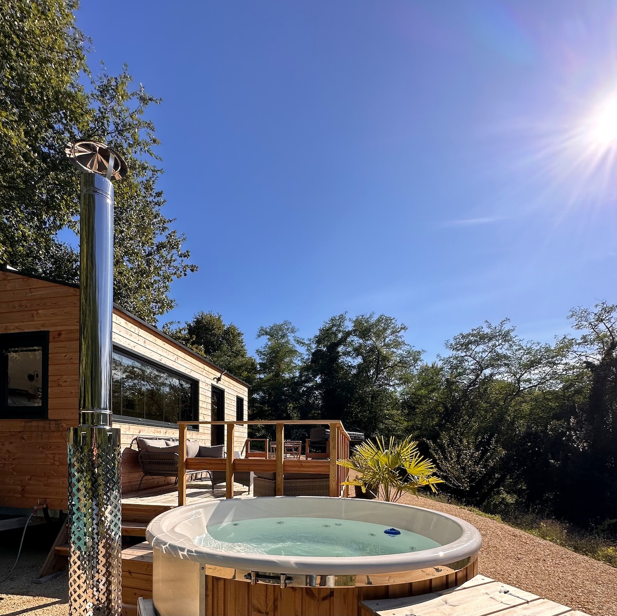 A private wooden spa is positioned on a spacious deck, surrounded by nature. A bright blue sky is visible, complemented by the warmth of sunlight. The Tiny House is seen in the background, and a comfortable seating area is accessible nearby.