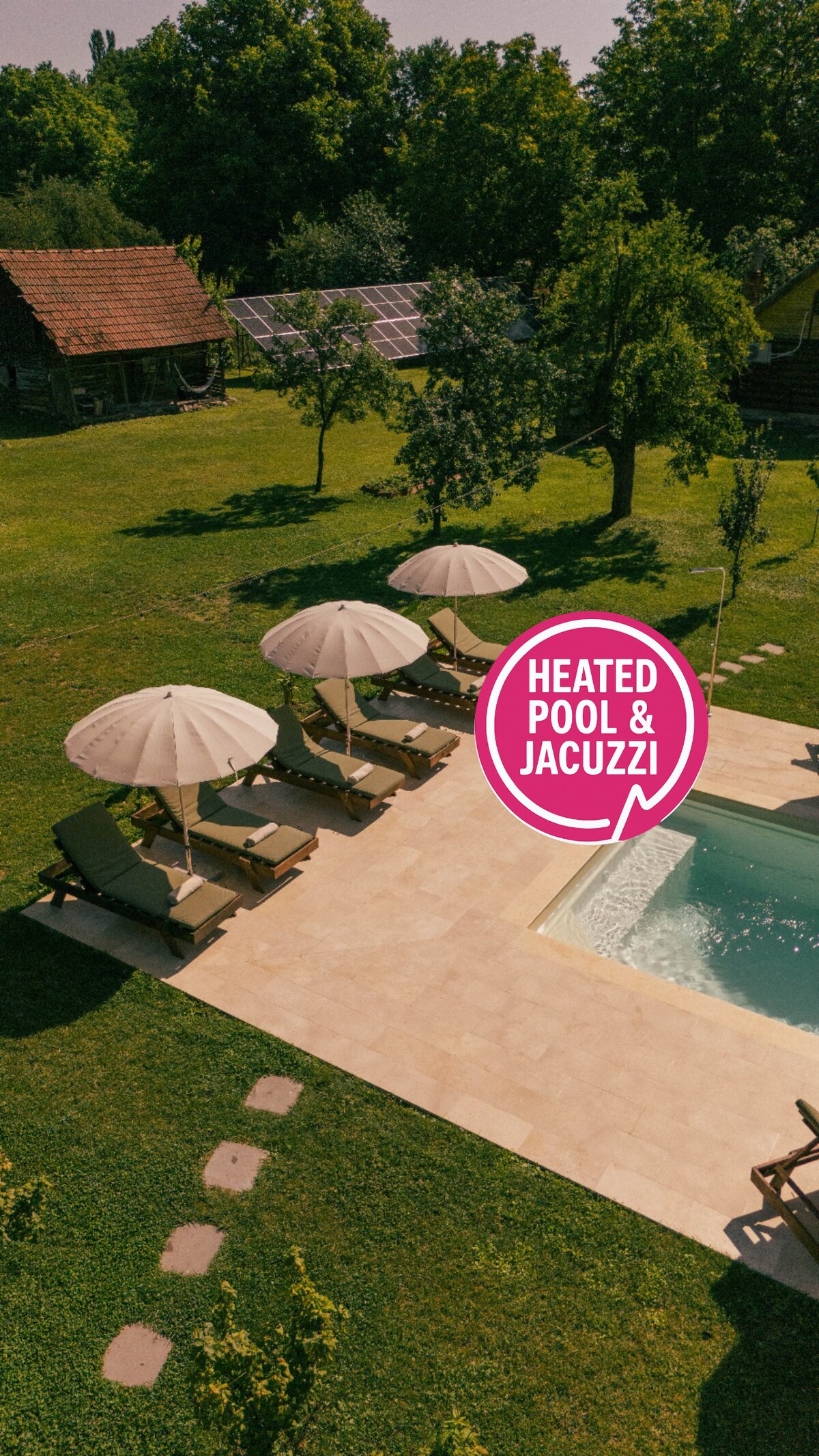 A spacious outdoor area showcases a heated swimming pool surrounded by several sun loungers and large umbrellas. Lush greenery and trees provide a serene backdrop, while a rustic building is visible in the distance, adding charm to the landscape.
