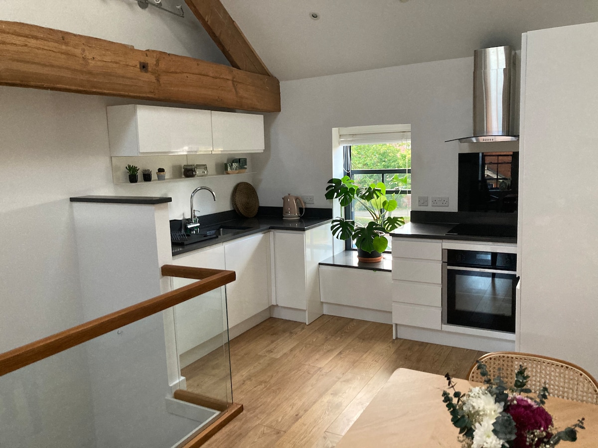 The kitchen features modern cabinetry and appliances with a black countertop. Natural light filters through a window, illuminating a potted plant on the counter. Exposed wooden beams add character to the airy space. A dining table is placed nearby, enhancing functionality.