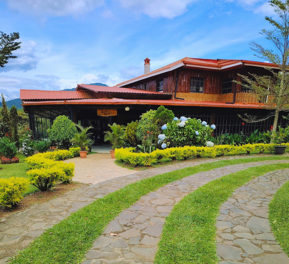 A spacious cabin is set amidst lush greenery, featuring a welcoming entrance lined with vibrant flowers and well-maintained shrubs. The wooden structure exudes warmth, complemented by the surrounding garden. A stone pathway leads visitors to the entrance, offering a glimpse of the inviting environment.