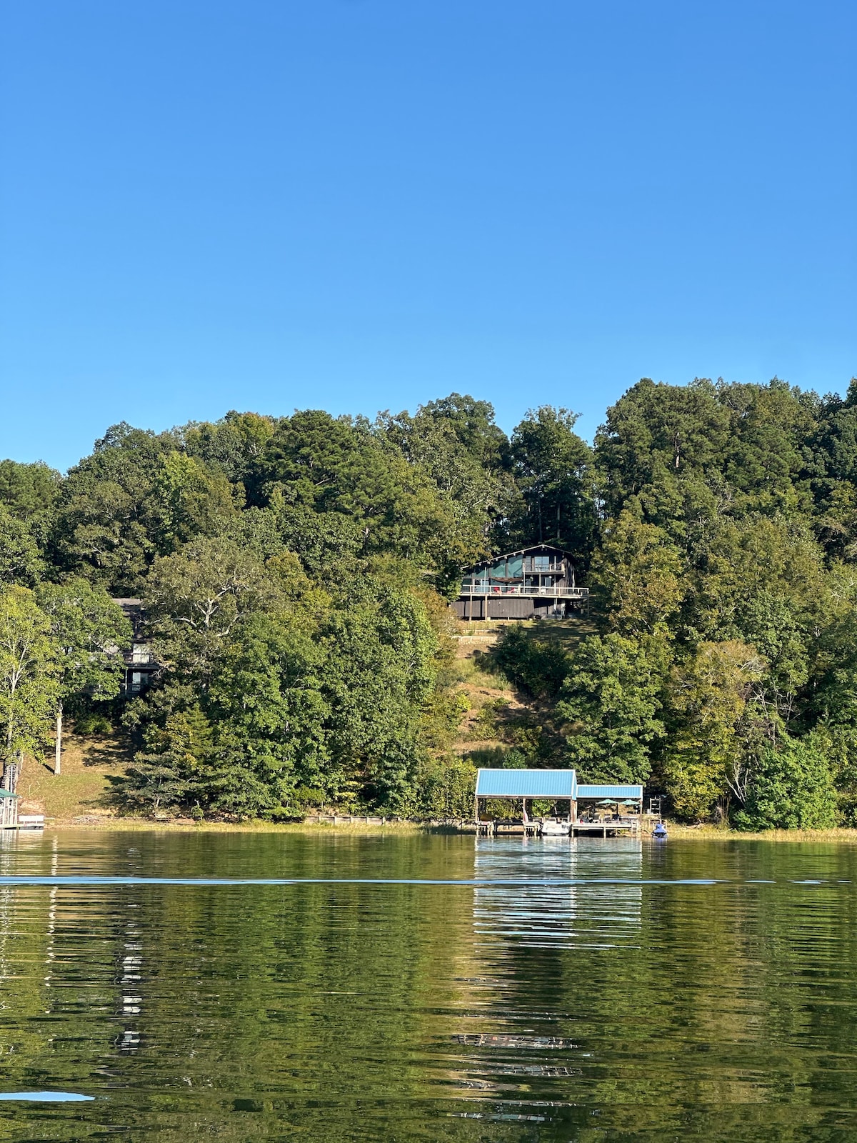 A serene view of a lakehouse perched among lush greenery, with a private dock visible at water's edge. The calm lake reflects the surrounding trees and blue sky, providing a peaceful atmosphere for relaxation.