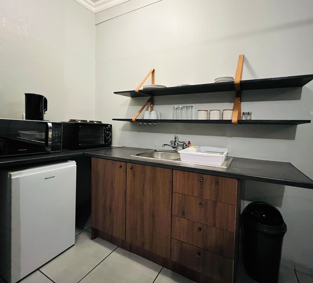 The open plan kitchen features a streamlined layout with modern appliances, including a microwave, refrigerator, and electric stove. Shelving displays glassware and dishware, while a sink is conveniently positioned. Underneath the counter, drawers and a waste bin offer practical storage solutions.