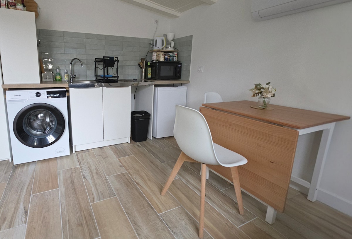 The kitchen area is equipped with modern appliances, including a washing machine, microwave, and refrigerator. A compact wooden table is accompanied by a white chair. Light-colored tiles line the floor, enhancing the openness of the space.