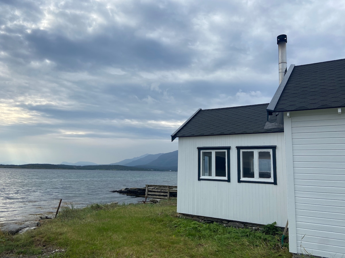 A traditional white cabin with a black roof is situated near the water's edge. The exterior features multiple windows framing views of the surrounding landscape. Grass extends towards the shoreline, where the mountains rise in the background under a cloudy sky.