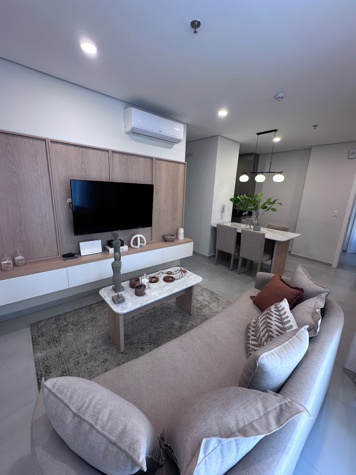 A modern and stylish living area is highlighted by a comfortable beige sofa adorned with decorative cushions. A neutral coffee table sits in front, with a flat-screen TV mounted on the wooden feature wall. Bright, overhead lighting illuminates the space, enhancing its contemporary aesthetic.