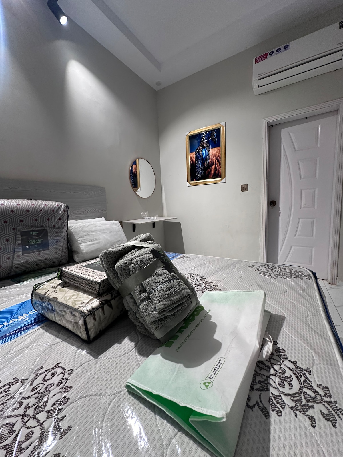 A well-lit sleeping area showcases a comfortable mattress with neatly arranged blankets and pillows. A circular mirror reflects the soft light, and a decorative framed artwork is mounted on the wall. A door leads to an adjoining space, enhancing the room's openness.