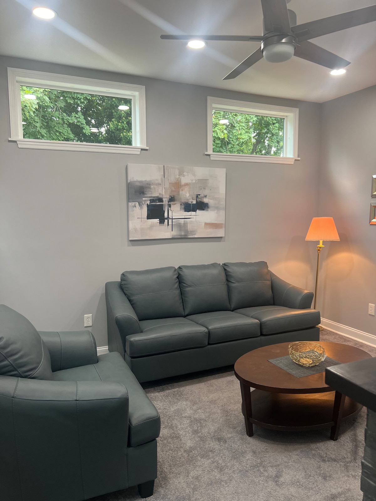A cozy living area is furnished with a dark leather sofa and a matching armchair. A round wooden coffee table sits in the center, topped with a decorative stone. Soft lighting from a nearby lamp and natural light from windows create a warm ambiance.