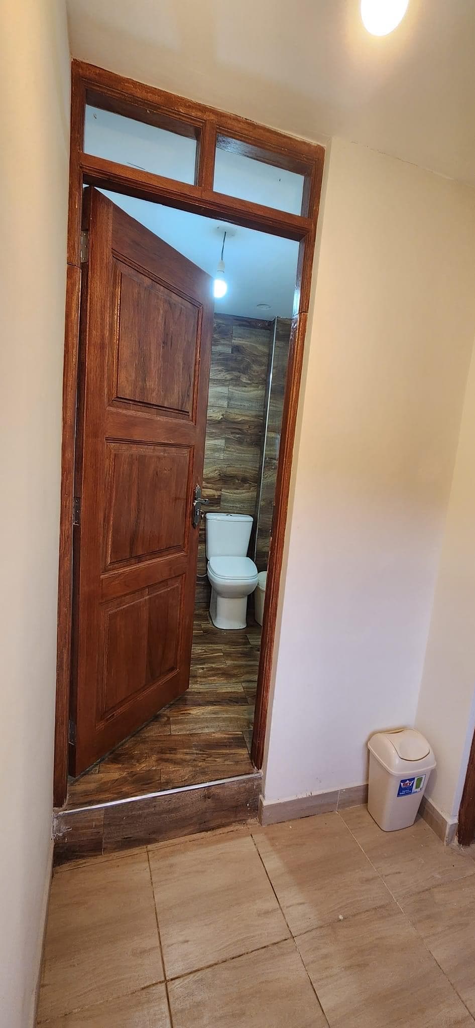 A wooden door is open, revealing a clean and modern bathroom with tiled walls. A toilet is visible, and warm lighting fixtures ensure adequate illumination. The hallway features tiled flooring that complements the bathroom's design, and a waste bin is placed in the corner.