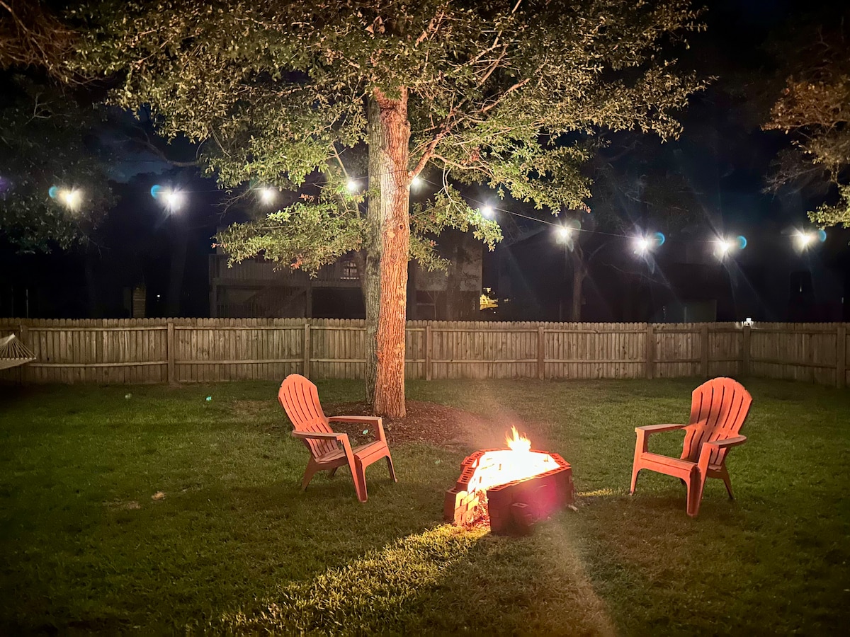 A cozy outdoor space is illuminated by string lights, creating a welcoming ambiance. Two wooden chairs surround a central fire pit, which emits a warm glow. The area features a grassy yard enclosed by a wooden fence and a tree providing gentle shade.