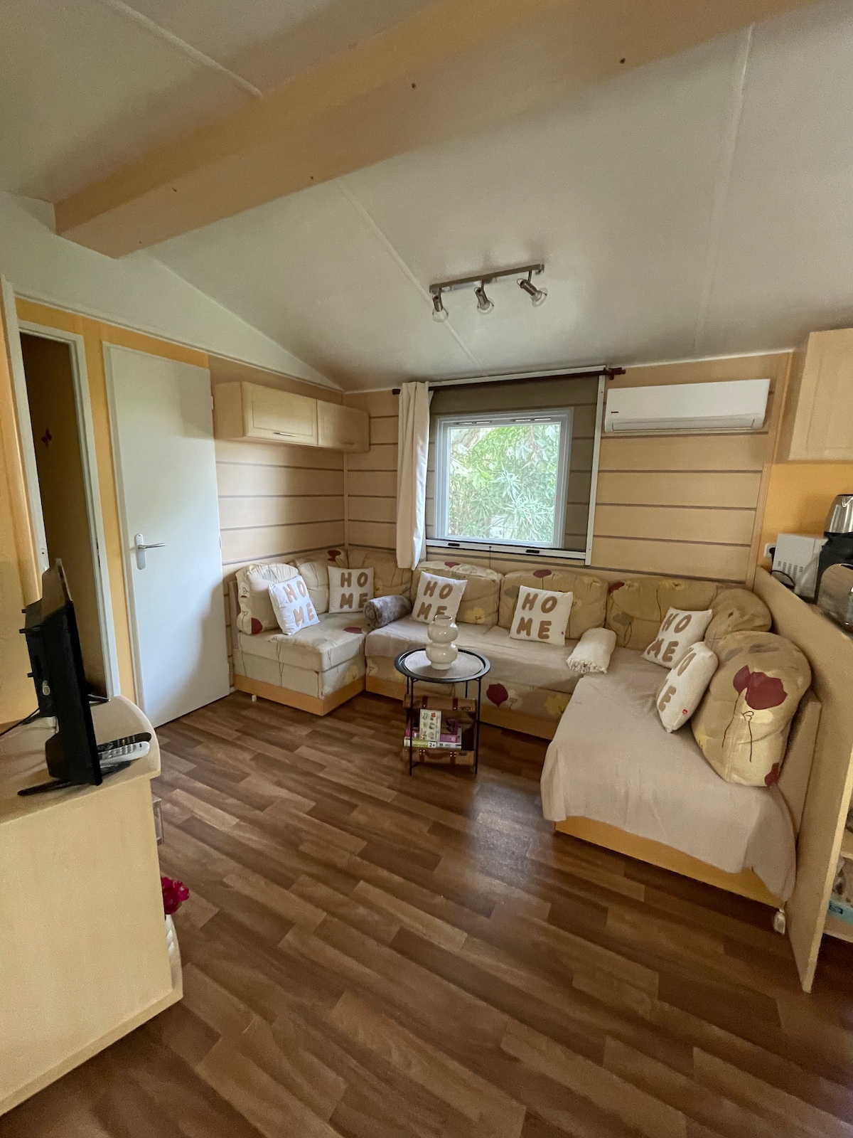 A cozy living area features a comfortable corner sofa adorned with decorative cushions. Natural light filters through a window, complemented by light wood paneling and warm flooring. A small round table is positioned between the sofa and a television unit, enhancing the inviting atmosphere.