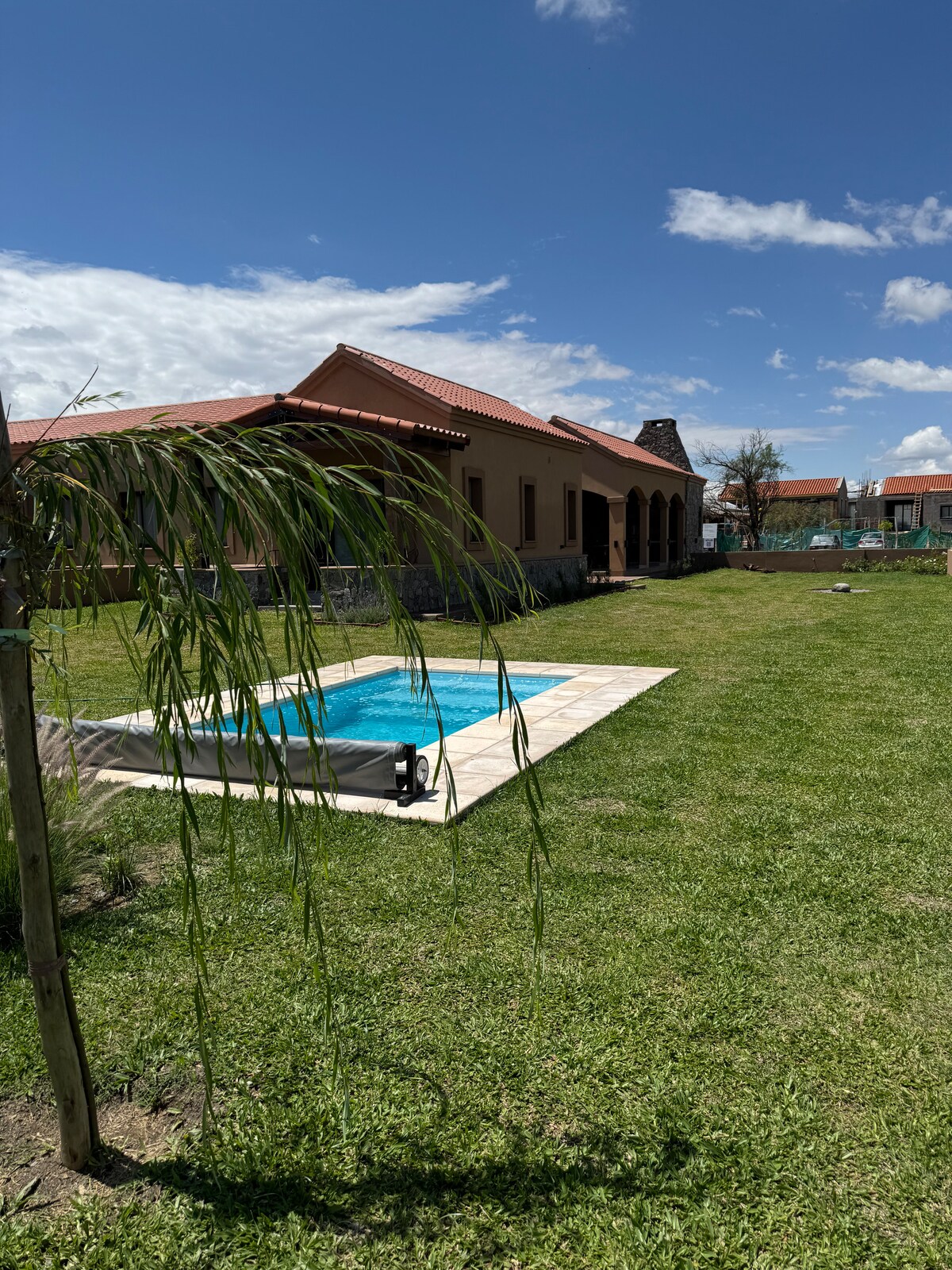 A well-kept yard features a rectangular swimming pool surrounded by lush green grass. The house, with a terracotta roof and arched porch, stands in the background under a clear blue sky, creating a serene outdoor space for relaxation.