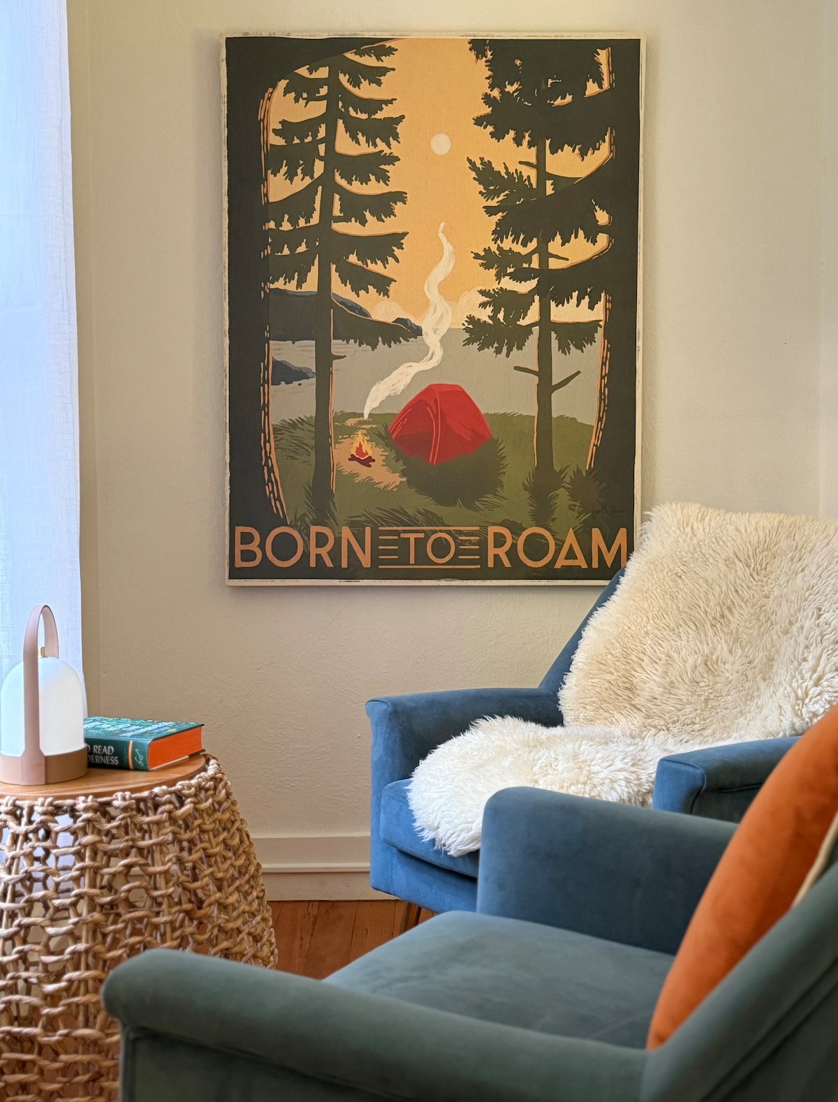 A wall-mounted artwork depicts a camping scene with a tent nestled among tall trees and a gentle smoke rising to the moon above. A cozy reading nook features a blue armchair with an orange accent pillow, alongside a woven basket table holding a soft throw and a book.