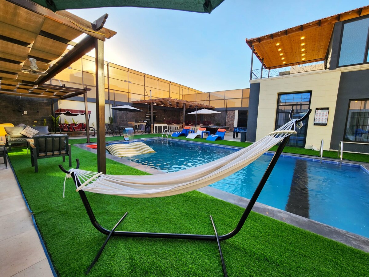 An outdoor area is depicted, featuring a pool surrounded by lounge chairs and a shaded hammock. A patio with stylish seating options and a shaded structure creates an inviting space, while green grass adds a touch of nature to the setting.